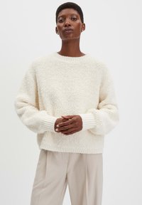 Dikke, crèmekleurige gebreide sweater met een gestructureerd oppervlak, geribbelde manchetten en een brede ronde hals. Gecombineerd met lichtbeige broek.