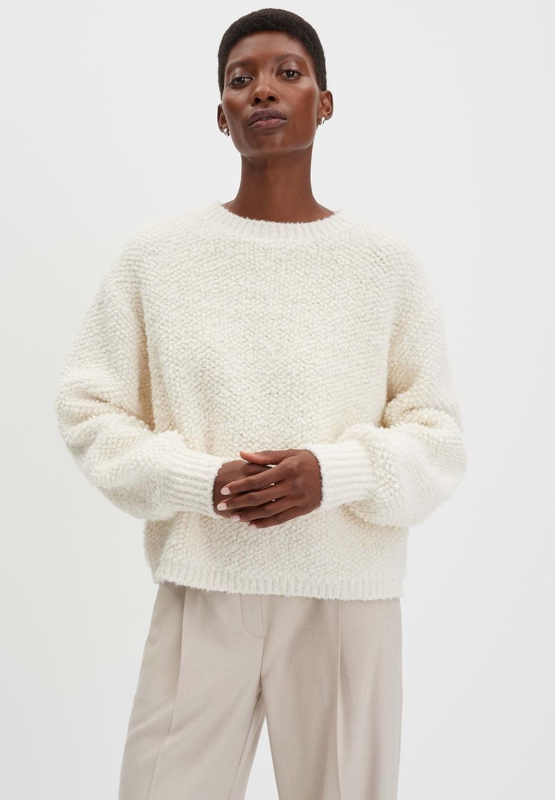 Dikke, crèmekleurige gebreide sweater met een gestructureerd oppervlak, geribbelde manchetten en een brede ronde hals. Gecombineerd met lichtbeige broek.