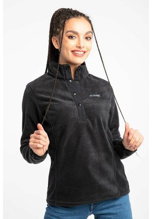 Uśmiechnięta kobieta z warkoczami, ubrana w czarny polar Columbia i niebieskie dżinsy, trzyma obiema rękami dwa warkocze.