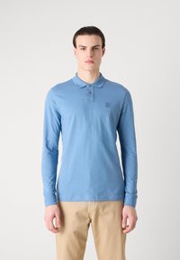Poloko s dlhým rukávom v oblohovomodrej farbe, vyrobené z mäkkého bavlneného materiálu. Má golier, tri gombíky a malé logo na ľavej hrudi.