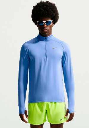 Uomo con afro che indossa occhiali da sole sportivi bianchi, maglietta azzurra chiaro a maniche lunghe con zip e pantaloncini Nike giallo neon, in piedi su sfondo bianco.