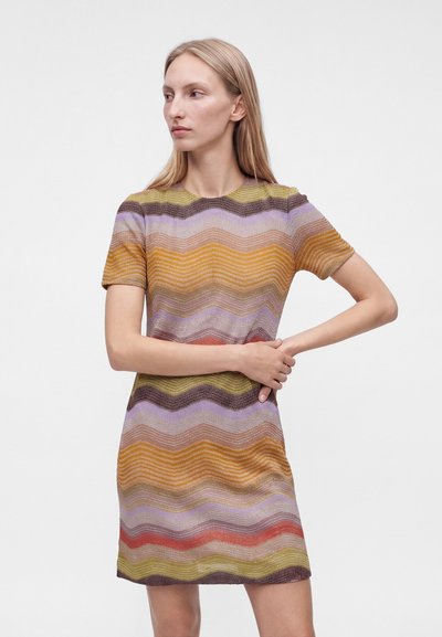 Femme aux longs cheveux blonds portant une robe à manches courtes avec des rayures ondulées horizontales multicolores, se tenant devant un fond blanc uni.