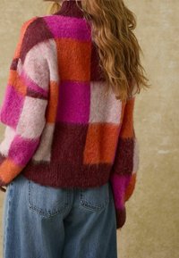 Bunter Wollmischpullover mit einem großen Karo-Muster in Pink, Orange und Burgunderrot sowie einem hohen Kragen. Getragen mit hellblauen Jeans.