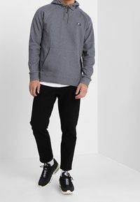 Grå munkjacka med dragsko och framficka, kombinerad med svarta byxor och svarta sportskor med vita detaljer.