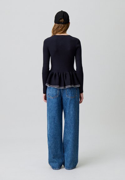 Donna in piedi con le spalle rivolte verso l'osservatore, indossa un berretto nero, una maglia peplum a maniche lunghe nera con bordi bianchi e jeans di denim blu larghi.