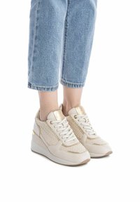 Zapatillas beige con paneles de punto texturizado, acentos dorados y cordones blancos. Cuenta con un collar acolchado y tacón cuña. Combinadas con vaqueros azul cortos.