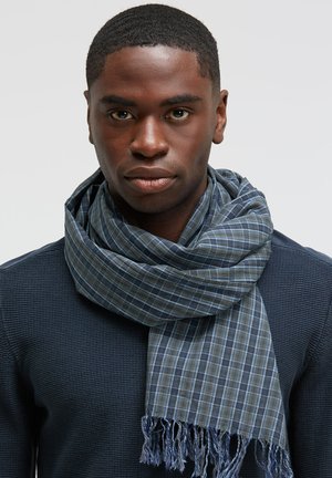 Blue plaid scarf made of soft fabric, featuring fringed edges. Worn loosely around the neck over a textured dark top.
