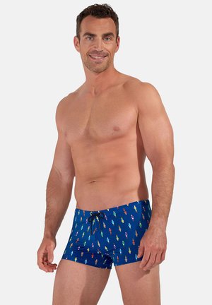 Smiling shirtless man wearing blue swim trunks with small colorful ice cream cone patterns and black drawstring, standing against plain background.