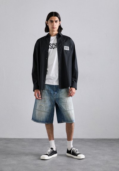 Camicia nera con bottoni e etichetta sul petto, indossata sopra una maglietta bianca con grafica. Combinata con pantaloni corti in denim chiaro e strappati e sneakers nere.