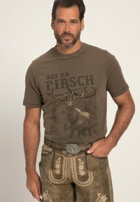 Camiseta gráfica marrón con diseño de ciervo y montaña, combinada con unos lederhosen beige con bordados florales y una hebilla de cinturón decorativa.