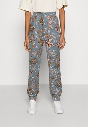 Osoba nosząca niebieskie spodnie z wysokim stanem z dużymi kwiatowymi i paisley wzorami, w połączeniu z białymi trampkami i kremową krótką bluzką na rękawach.