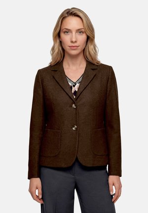 Femme aux cheveux blonds portant un blazer marron foncé à deux boutons sur un haut à motifs et un pantalon sombre, debout devant un fond uni.
