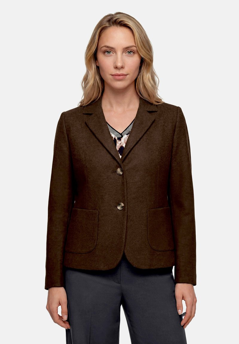 Femme aux cheveux blonds portant un blazer marron foncé à deux boutons sur un haut à motifs et un pantalon sombre, debout devant un fond uni.