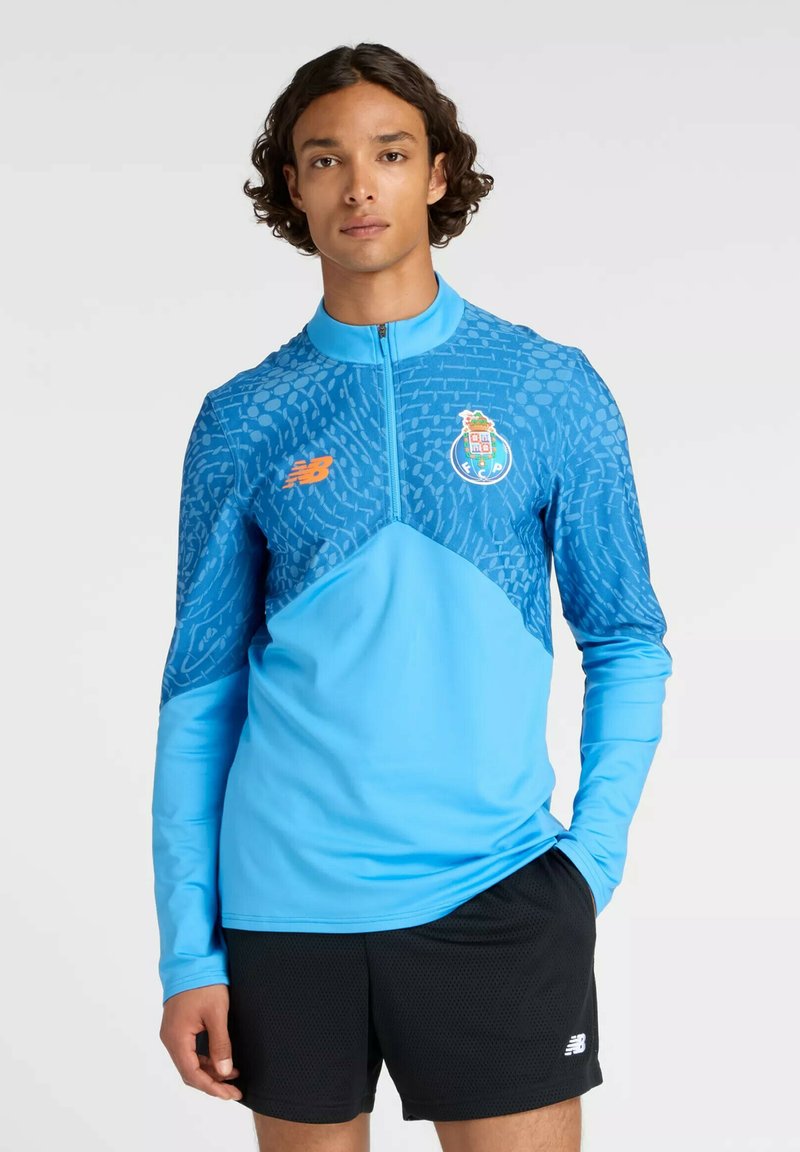 Jonge man die een blauw sportshirt met patroon en rits draagt, samen met zwarte sportshorts, staand met één hand in zijn zak.