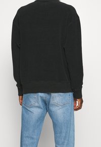Persona che indossa una maglietta nera a maniche lunghe e jeans in denim azzurri chiari, mostrata di spalle su uno sfondo bianco semplice.