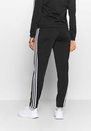Personne portant un pantalon de sport noir avec trois bandes blanches sur le côté, un haut à manches longues noir, des chaussettes grises et des baskets blanches, debout, dos tourné.