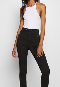 Pantalones skinny de cintura alta negros con bolsillos frontales con cremallera y un acabado de textura suave, combinados con una blusa halter blanca.