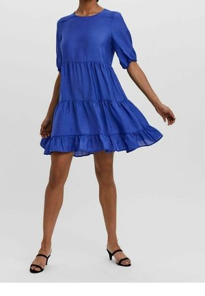 Robe bleue à volants avec des manches courtes bouffantes, fabriquée en tissu texturé, dotée d'un col rond et d'un ourlet fluide. Des escarpins noirs à lanières sont portés.