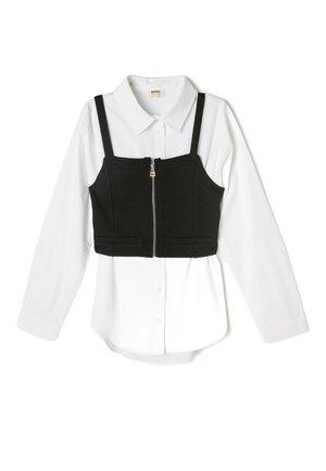 Schwarzes, kurzes Top mit verstellbaren Trägern über einem weißen Langarmshirt. Hat einen Reißverschluss vorne und zwei Taschen auf der Vorderseite.