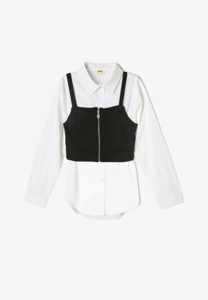 Sort cropped top med justerbare stropper over en hvid langærmet skjorte. Har en frontlynlås og to lommer foran.