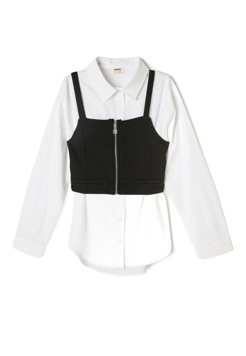 Top negro corto con tirantes ajustables sobre una camiseta blanca de manga larga. Cuenta con una cremallera frontal y dos bolsillos en la parte superior.