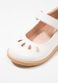 Białe lakierowane buty typu Mary Jane z wycięciami na palcach, brązową gumową podeszwą i regulowanym paskiem. Gładkie wykończenie i wyściełane wnętrze.