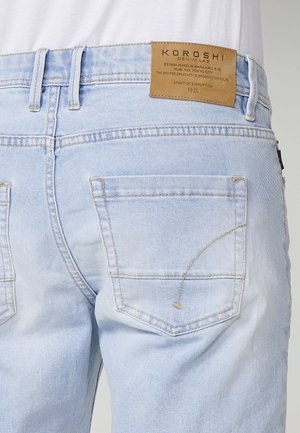 Jeans in denim blu chiaro con una patch in pelle tan con logo e testo. Dettagli sfilacciati e doppie tasche posteriori con cuciture gialle.