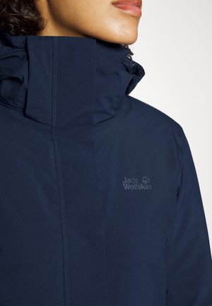 Kvinde iført en marineblå Jack Wolfskin jakke med høj krave, der viser mærkets logo på brystet mod en lys baggrund.