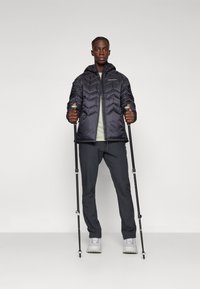 Schwarze gepolsterte Jacke mit Zickzackmuster, Kapuze und einem Frontreißverschluss, getragen mit grauen Hosen und grauen Wanderschuhen, während zwei Trekkingstöcke gehalten werden.