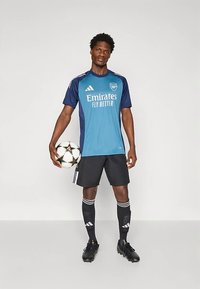 Sininen ja laivastonsininen jalkapallopaita, jossa on "Emirates Fly Better" -logo, mustat shortsit, mustat nappikset ja polveen ulottuvat raidalliset sukat, pitäen jalkapalloa.