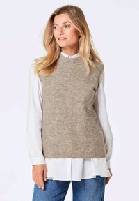 Pull en tricot sans manches de couleur brun clair avec une texture côtelée. Superposé sur une chemise blanche à col, présentant une coupe décontractée et un col rond.