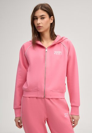 Jonge vrouw die een roze hoodie met ritssluiting en bijpassende joggingbroek draagt, beide met witte "JOOP! JEANS" logo's.