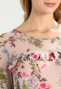 Blusa floral con una tela transparente de color rosa claro y estampados de flores coloridos, un escote redondo y delicados acentos de volantes a lo largo de la costura.