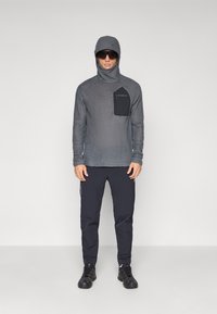 Sporty grå langermet hettegenser med teksturert stoff og en svart brystlomme. Kombinert med mørk marineblå bukser og svarte fottøy.