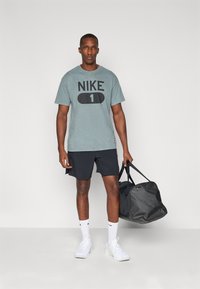Grå Nike t-shirt med "NIKE 1" trykt i fed sort, parret med mørke shorts. Modellen holder en sort duffeltaske, iført hvide sneakers og sokker.