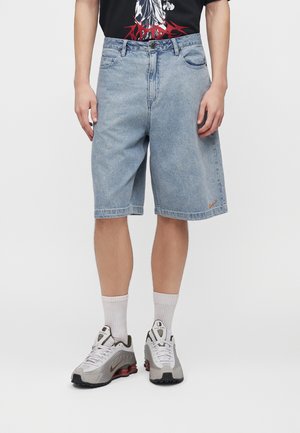 Persoon draagt lichtblauwe denim knielange short met klein oranje logo, witte sportsokken en witte, grijze en rode sneakers, staand op een witte achtergrond.