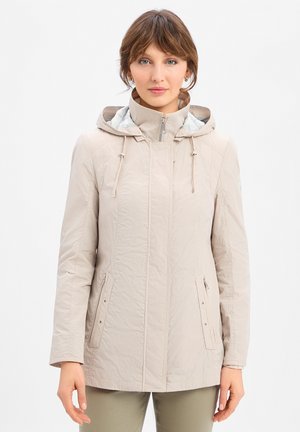 Vrouw die een beige capuchonjas met rits en trekkoord draagt, staand en vooruitkijkend tegen een effen witte achtergrond.