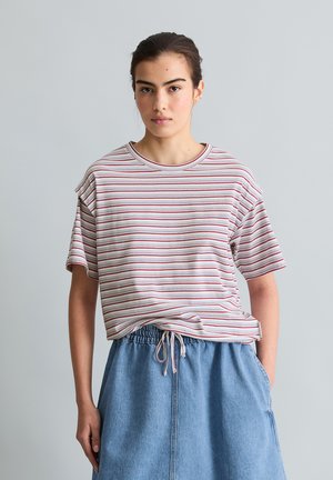 Jonge vrouw staat tegen een grijze achtergrond, draagt een gestreept t-shirt en een blauwe denim rok met een taillekoord, handen in de zakken.