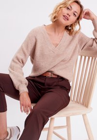 Lys beige sweater i blødt strik med V-hals, parret med brune skræddersyede bukser. Outfitten har en mønstret, rund bæltesdetalje.