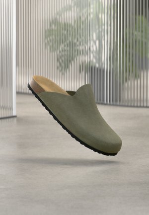 Olivengrøn ruskind slip-on mule med korksål og sort bund, svævende over en betonflade med slørede planter i baggrunden.