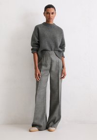 Grauer Strickpullover mit geripptem Ausschnitt, kombiniert mit hochtaillierten, weiten Karohosen. Model steht vor einem schlichten Hintergrund.