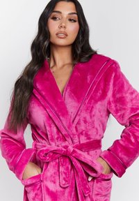 Accappatoio in spugna fucsia con collo a scialle, vita con cintura e tasche laterali. Caratterizzato da una superficie morbida e una leggera lucentezza.