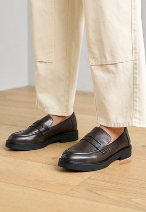 Brune læderloafers med en glat tekstur, med en dekorativ rem og en lav sort sål, kombineret med lyse bukser.