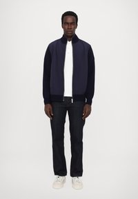 Navy bomber jacket with ribbed knit collar and cuffs, paired with a white polo and dark jeans. Shoes are white with laces.