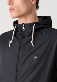 Sort letvægtsjakke med hætte, der har en frontlynlås, beige snoretræk og et rundt logo-patch på brystet.