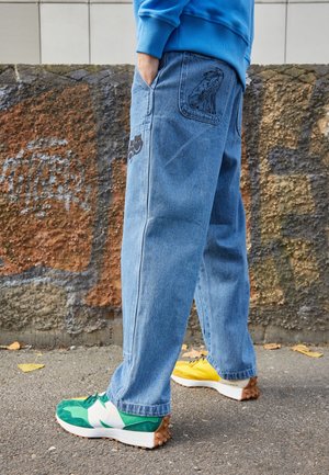 Persona con vaqueros azules con parches de perro y máscara de teatro, zapatillas verdes y amarillas, de pie sobre asfalto cerca de una pared texturizada con grafiti.