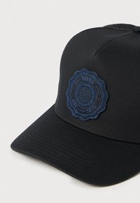 Μαύρο καπέλο με δομημένο σχεδιασμό, που διαθέτει υφασμένο μπλε σκούρο έμπλαστρο με ανάγλυφο λογότυπο. Η γείσος είναι επίπεδη, με λεπτή ραβδωτή λεπτομέρεια.