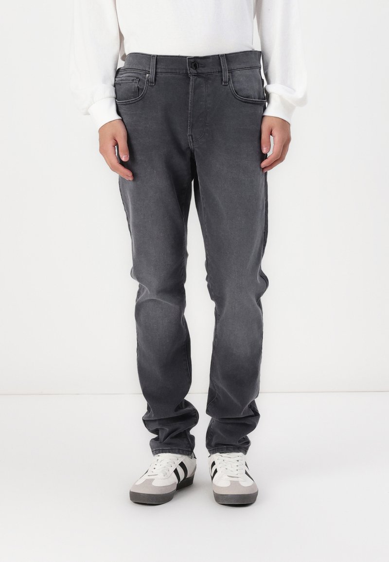 Szürke farmernadrág, slim fit szabással, öt zsebbel, enyhe kopással és standard gombos záródással, fehér sportcipővel párosítva.