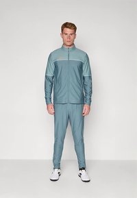 Vīrietis stāv, ģērbies gaiši zilā un zaļgani zilā sporta tērpā ar baltām sporta apaviem, pret vienmērīgu balto fonu.