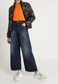 Puchowa kurtka bomber w kamuflażu, pomarańczowa koszulka z okrągłym dekoltem, szerokie ciemnoniebieskie dżinsy oraz masywne białe sneakersy.